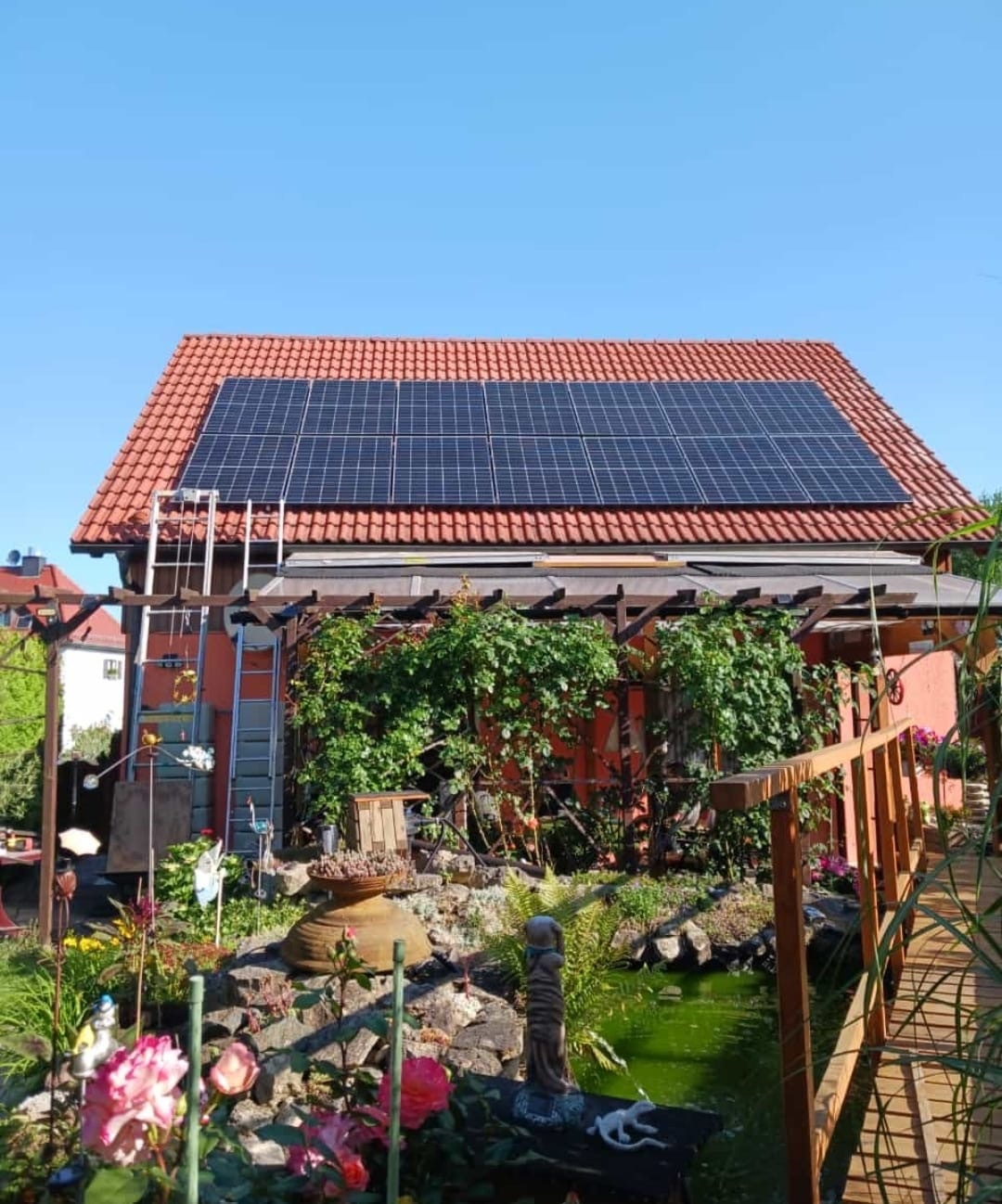 Eine Haus mit roten Dachziegeln und Solarpanelen, umgeben von Pflanzen und Teich.
