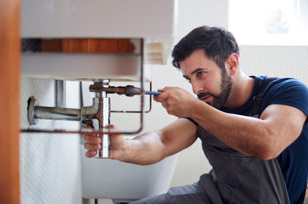 Male Plumber Using Wrench To Fix Leaking Sink In Home Bathroom
