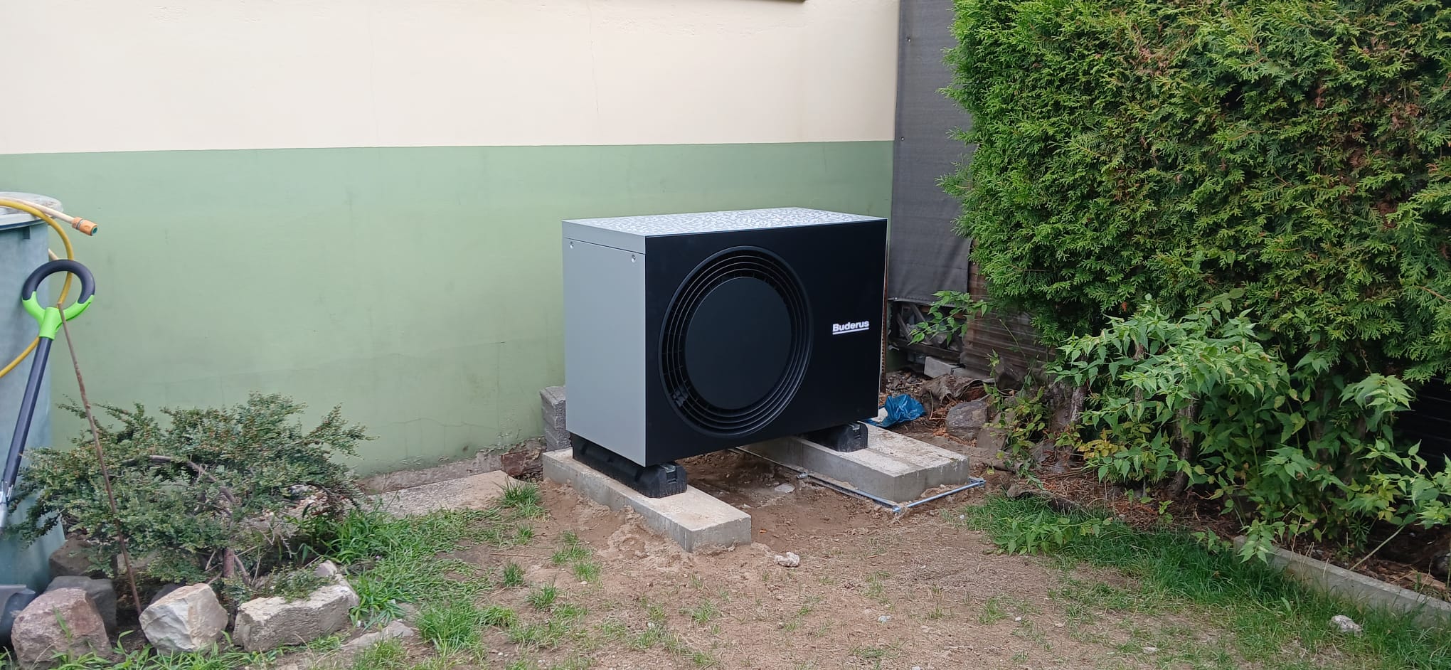 Eine schwarze Klimaanlage steht im Garten, umgeben von Büschen und Steinplatten.